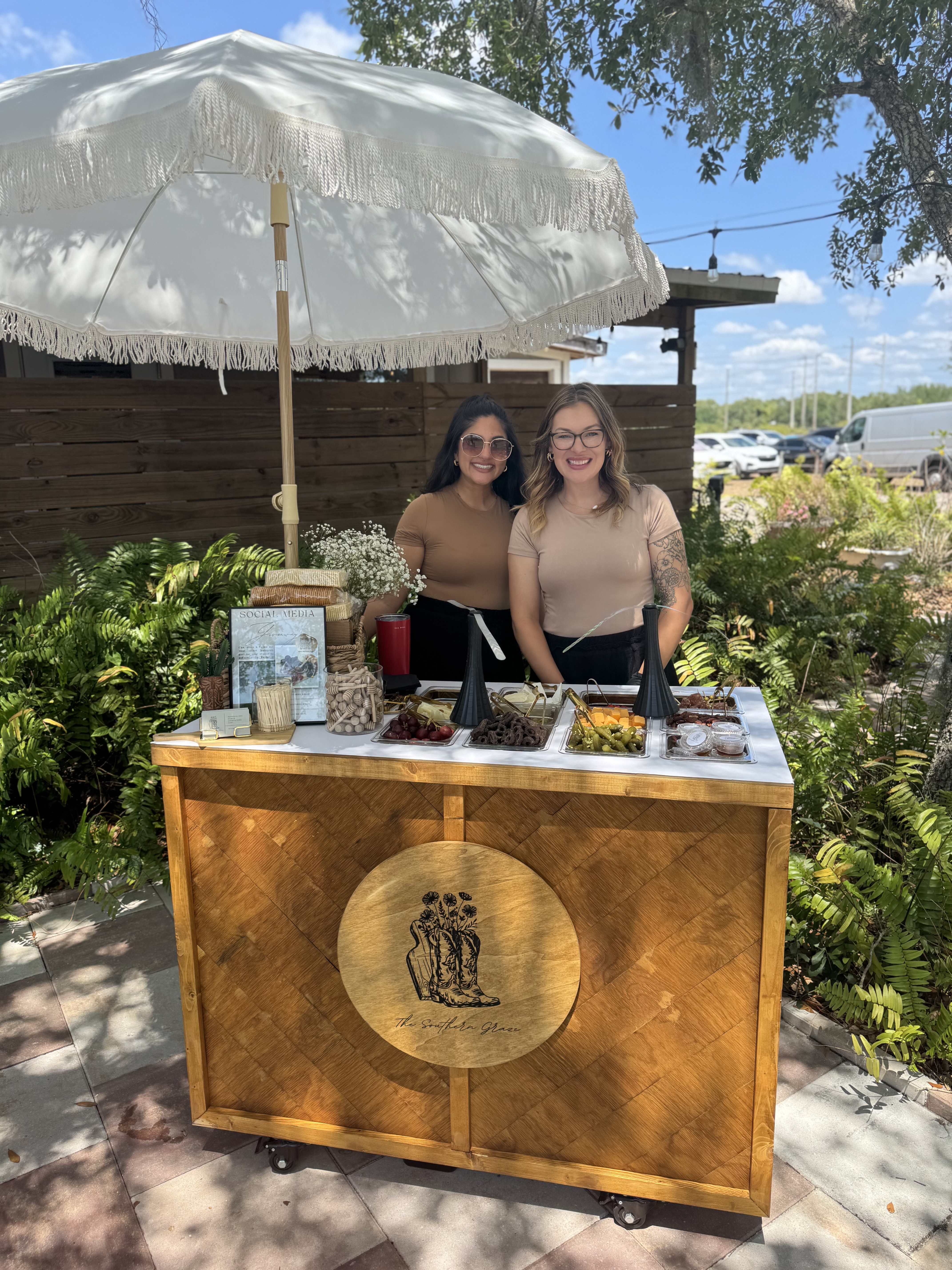 Two women smiling at the Southern Graze mobile charcuterie cart with umbrella